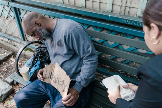 A person offers food to a man sitting on a park bench holding a 