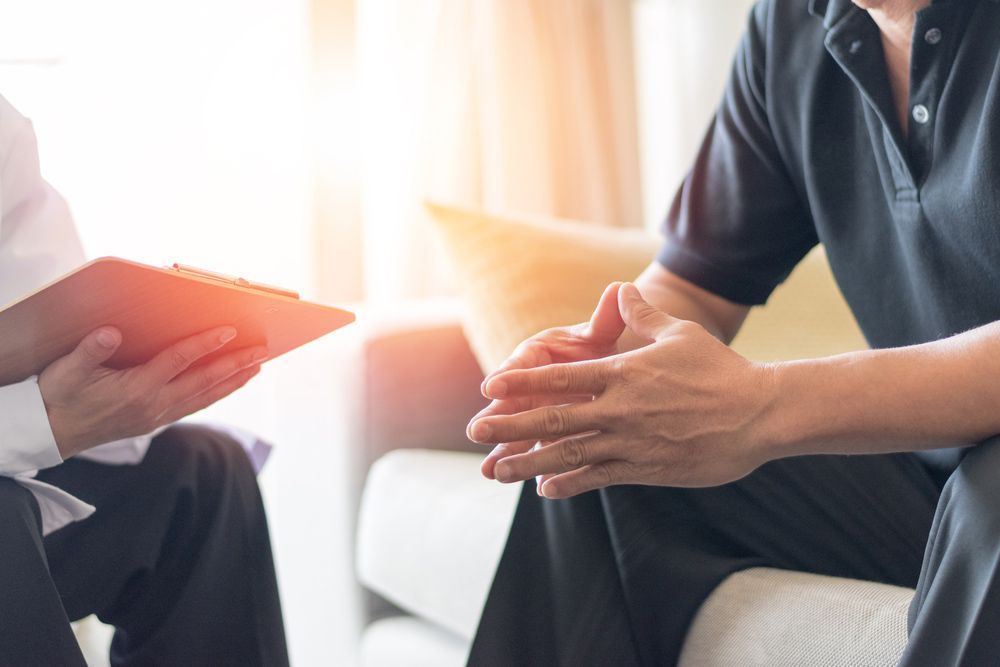 A Man is Sitting on a Couch Talking to a Doctor Who is Holding a Clipboard — One Stop Medical in Mackay, QLD