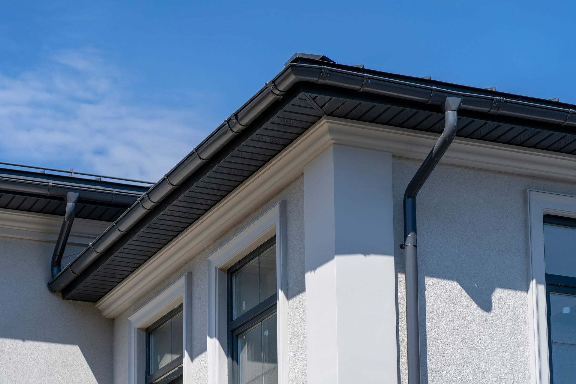 Corner of a light-colored house with dark roof, gutters, and trim against a blue sky.