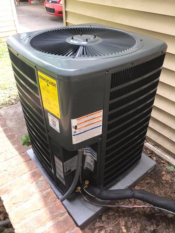 An outdoor air conditioning unit with a fan and black and grey exterior. It sits on a concrete pad near a building.