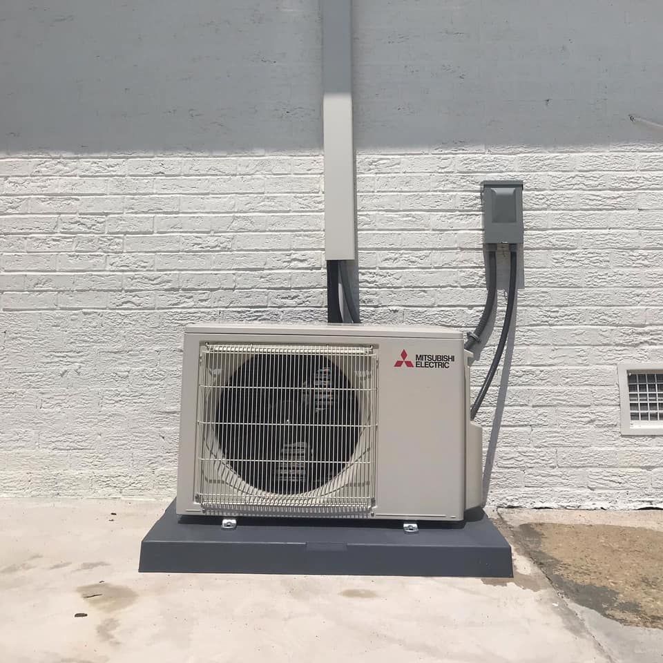 Mitsubishi air conditioning unit mounted on a concrete pad against a white brick wall.