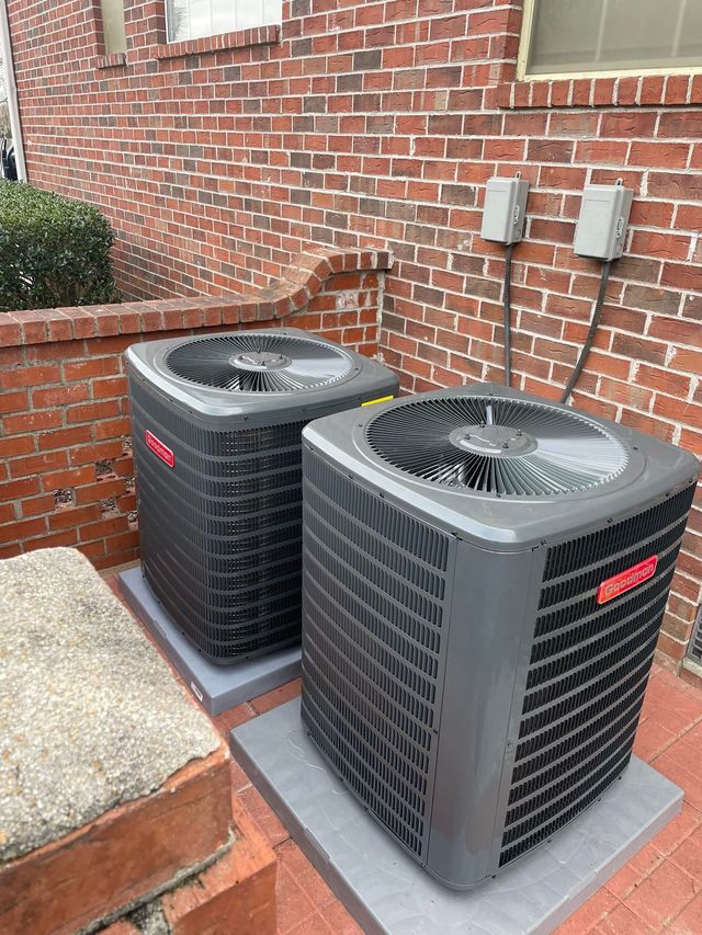Two Goodman air conditioning units on concrete pads against a brick wall, with electrical boxes above.