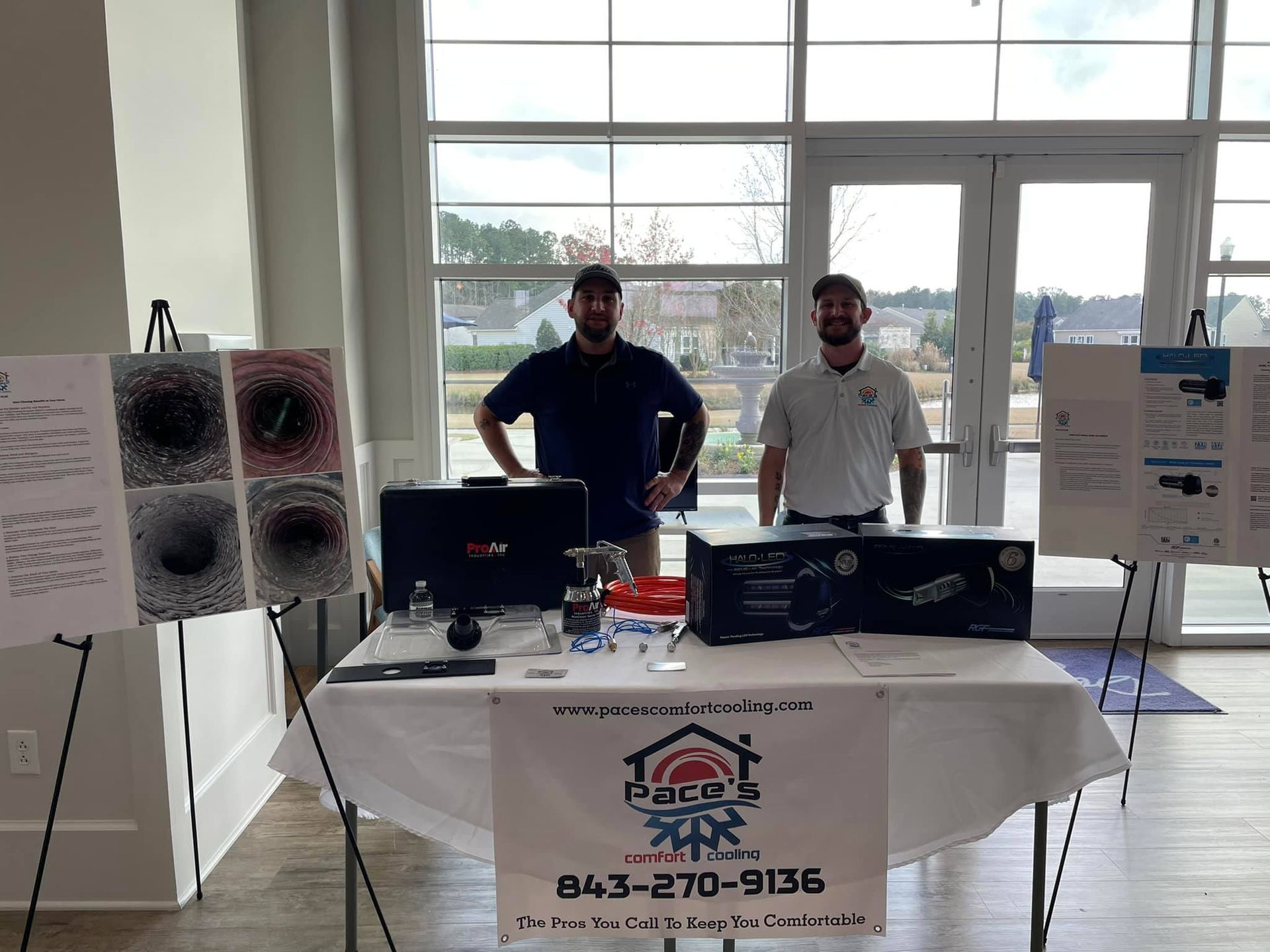 Two men at a vendor table with equipment, company logo, and contact info, inside a building with a view outside.
