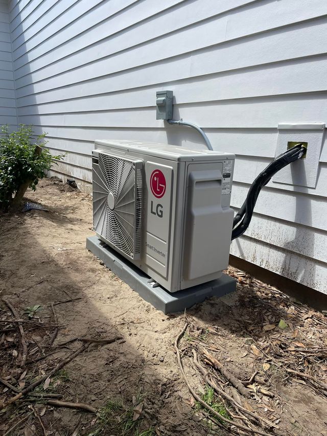 LG outdoor air conditioner unit installed on a concrete pad near a white house wall. Black power cables visible.
