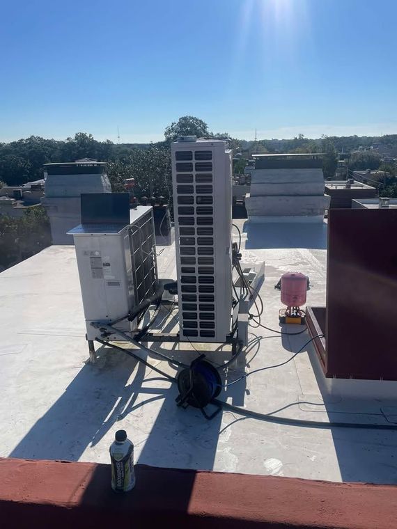 Rooftop HVAC unit with tall condenser, metal framework, and surrounding chimneys under a bright blue sky.