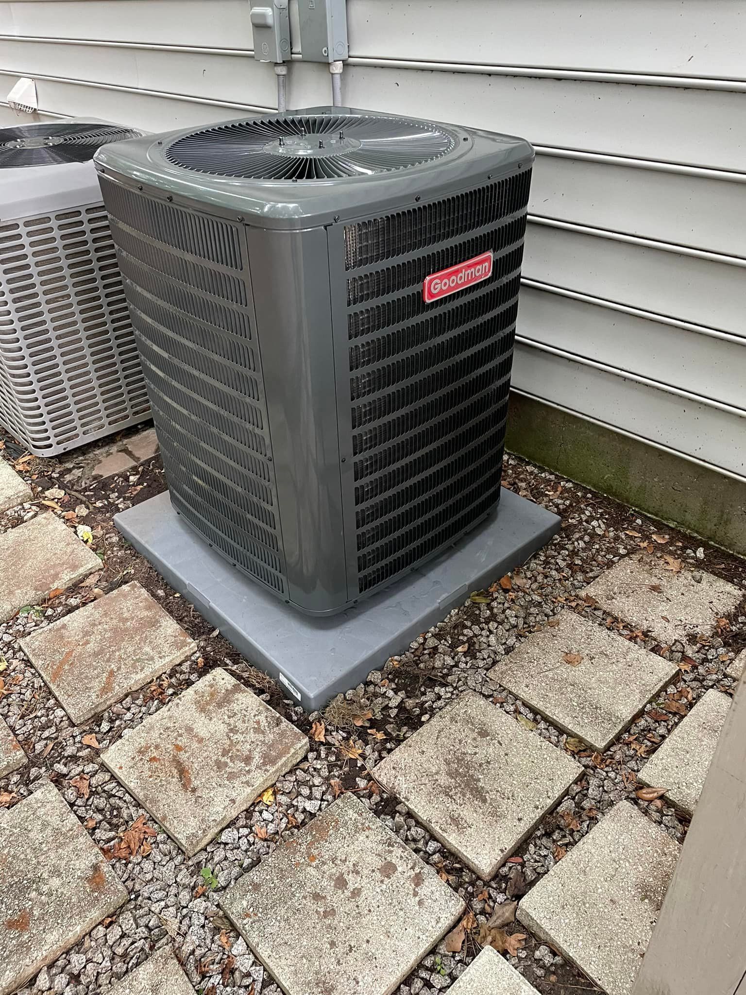 Outdoor Goodman air conditioning unit on a concrete pad, surrounded by stone pavers, against a white-sided building.
