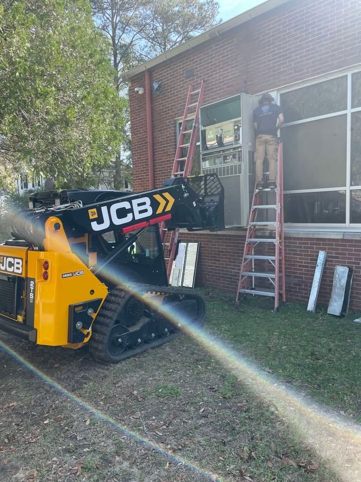 JCB compact track loader by a brick building with a worker on a ladder, replacing window frames.