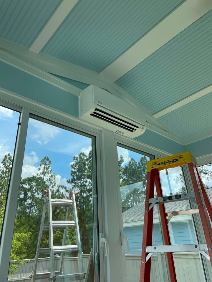 Air conditioner unit installed on a blue ceiling in a sunroom with windows and two ladders.