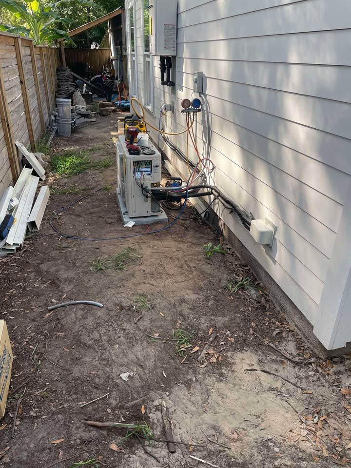 HVAC unit and piping against a white house wall in a dirt yard with a wooden fence.