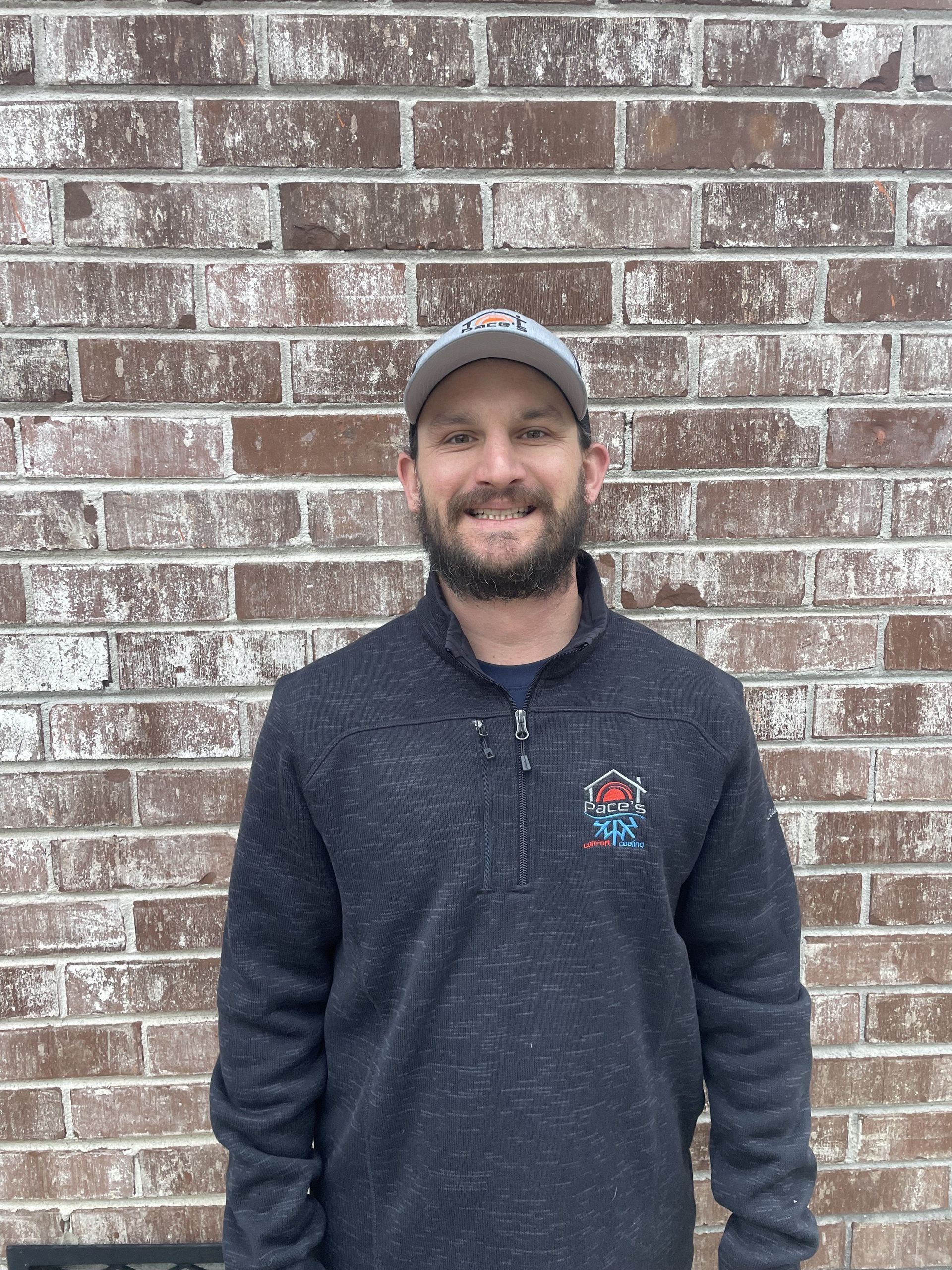 Man in a navy pullover and baseball cap smiles in front of a brick wall.