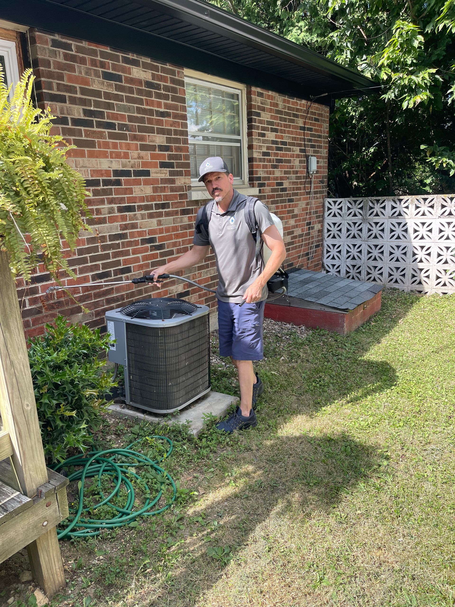 Man spraying insecticide near an AC unit outside a brick house.
