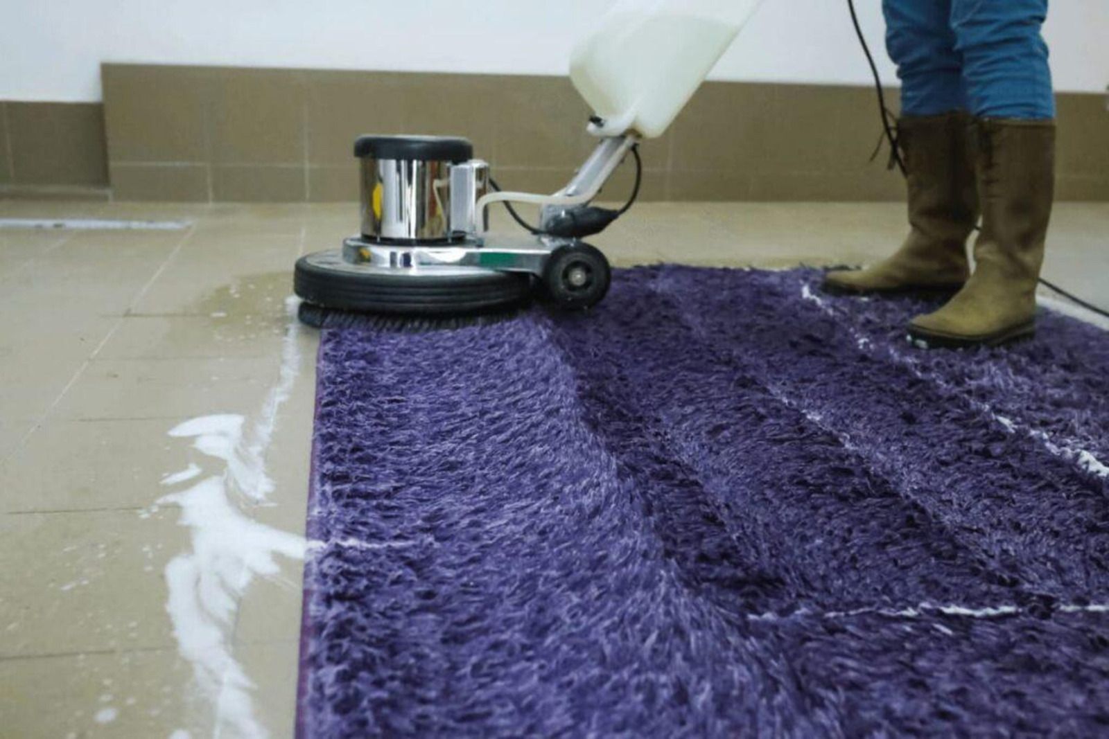 Person cleaning a purple rug with a floor buffer in a tiled room.