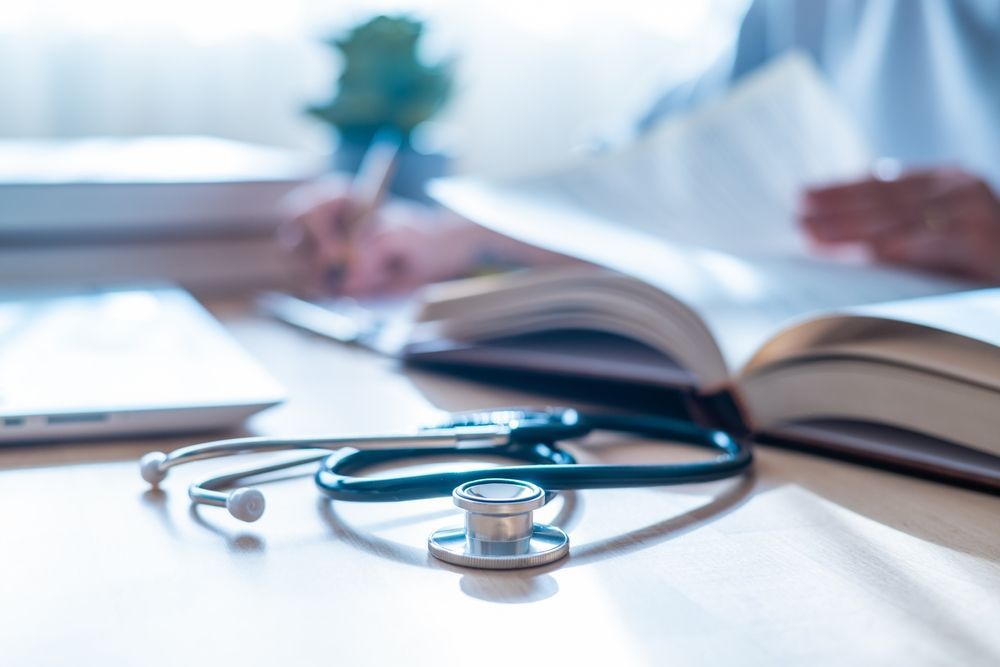 A stethoscope rests on a desk next to an open book and a blurred person writing notes.