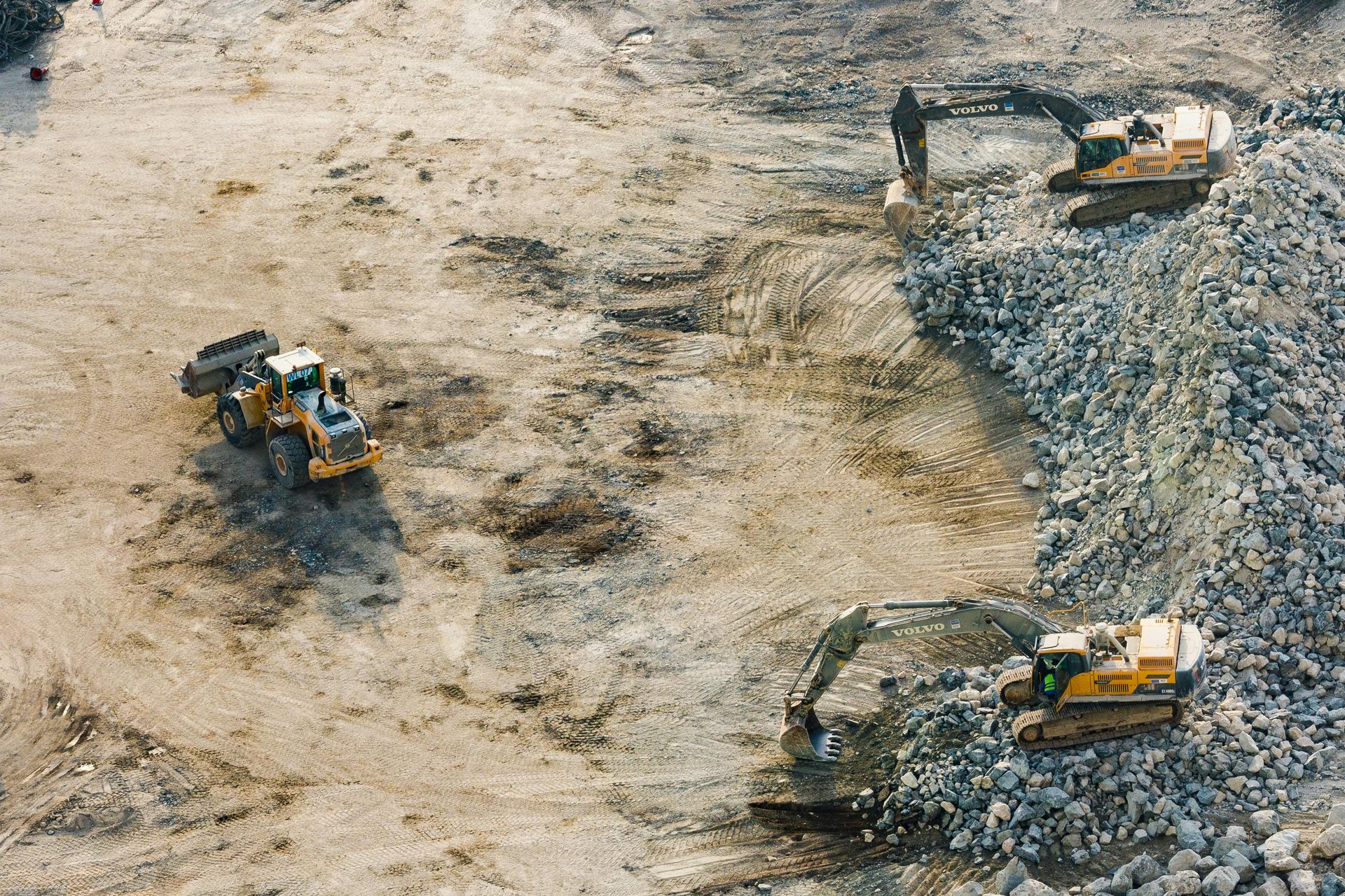 Three yellow construction vehicles operate on a dirt and gravel site, moving rocks and leveling the ground.
