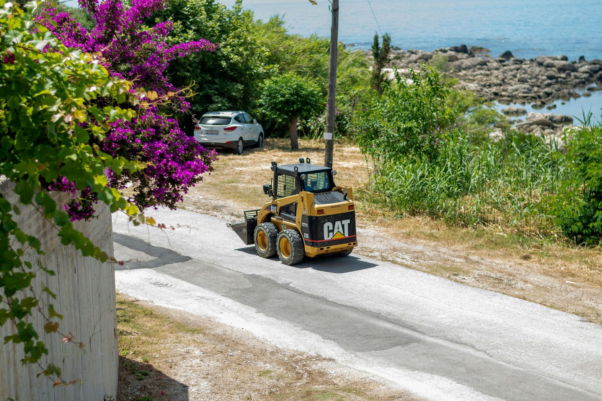 A yellow CAT skid-steer loader parked on a gravel path near a beach, with vibrant purple flowers in the foreground.