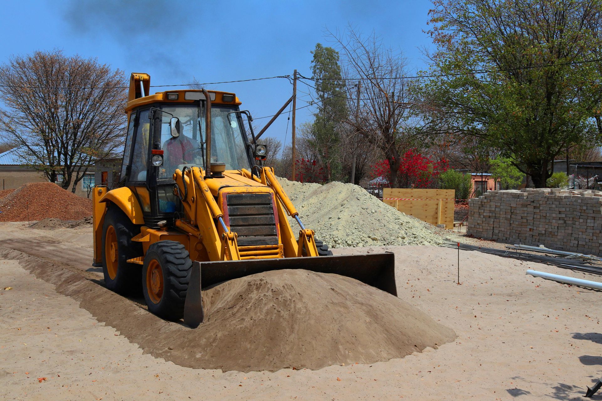 A yellow backhoe loader pushes a mound of sand at a construction site on a sunny day.