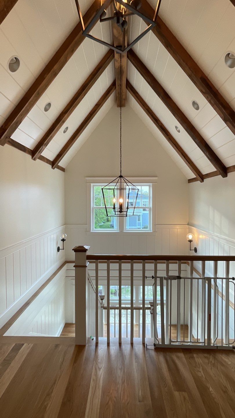 Interior view of a light-filled hallway with a high, sloped ceiling, wooden beams, and a chandelier.  A balcony overlooks the scene.