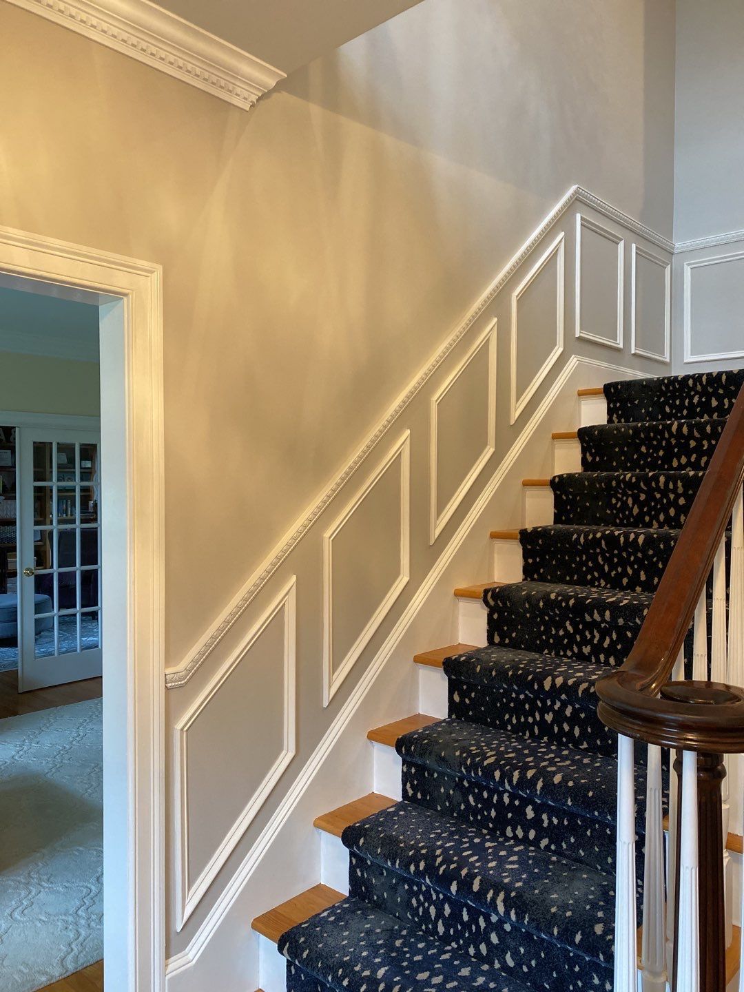A staircase with a dark blue carpet, wooden handrail, and beige walls with white decorative trim. The door frame to the left has similar trim.