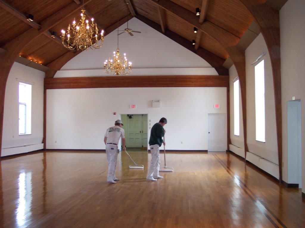 Three people refinish a wooden floor in a large room with a vaulted ceiling, chandeliers, and tall windows.
