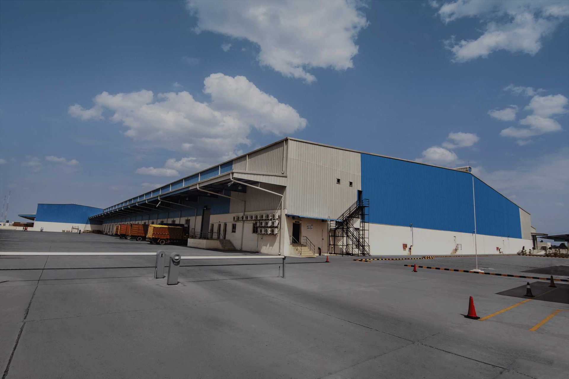 Large blue and white warehouse with loading docks, under a partly cloudy sky. A metal gate is in the foreground.