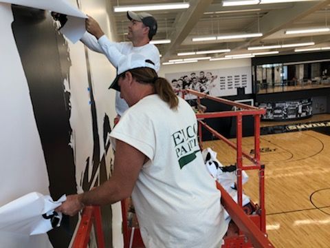 Two people installing a large black and white wall graphic on a wall in a gymnasium. One person is on a lift.