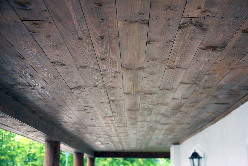 Wooden ceiling boards, stained brown, forming a covered porch ceiling. The view is from below, with the porch structure and greenery visible.