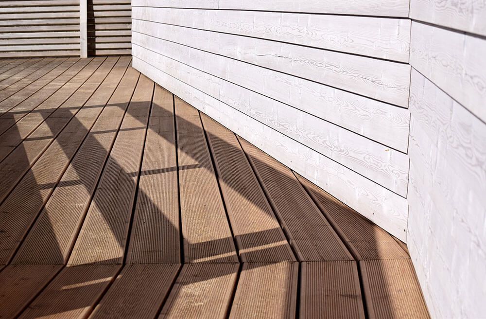 Wooden deck with brown planks, shadowed by a white slatted structure. Bright sunlight creates contrast on the surfaces.