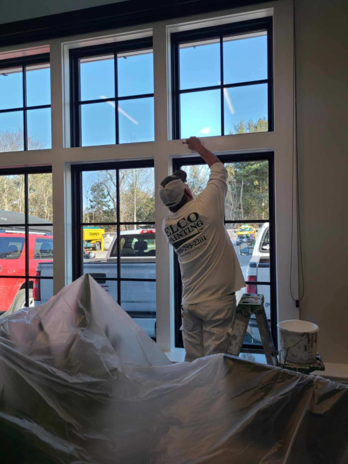 A painter in white overalls and cap is painting a white window frame. The windows are black-framed and the setting is indoors, overlooking vehicles and trees.