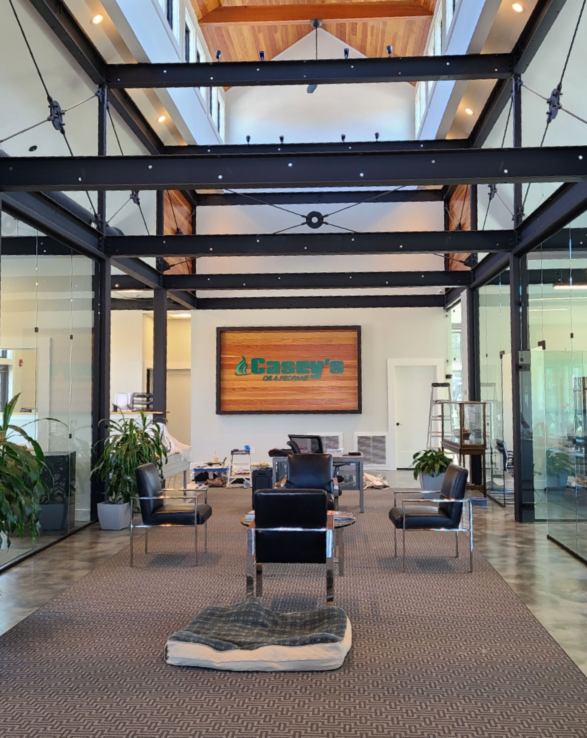 Modern waiting area with a large, wooden sign. Black beams frame glass walls. Chairs are arranged around a stone-like dog bed on a rug.