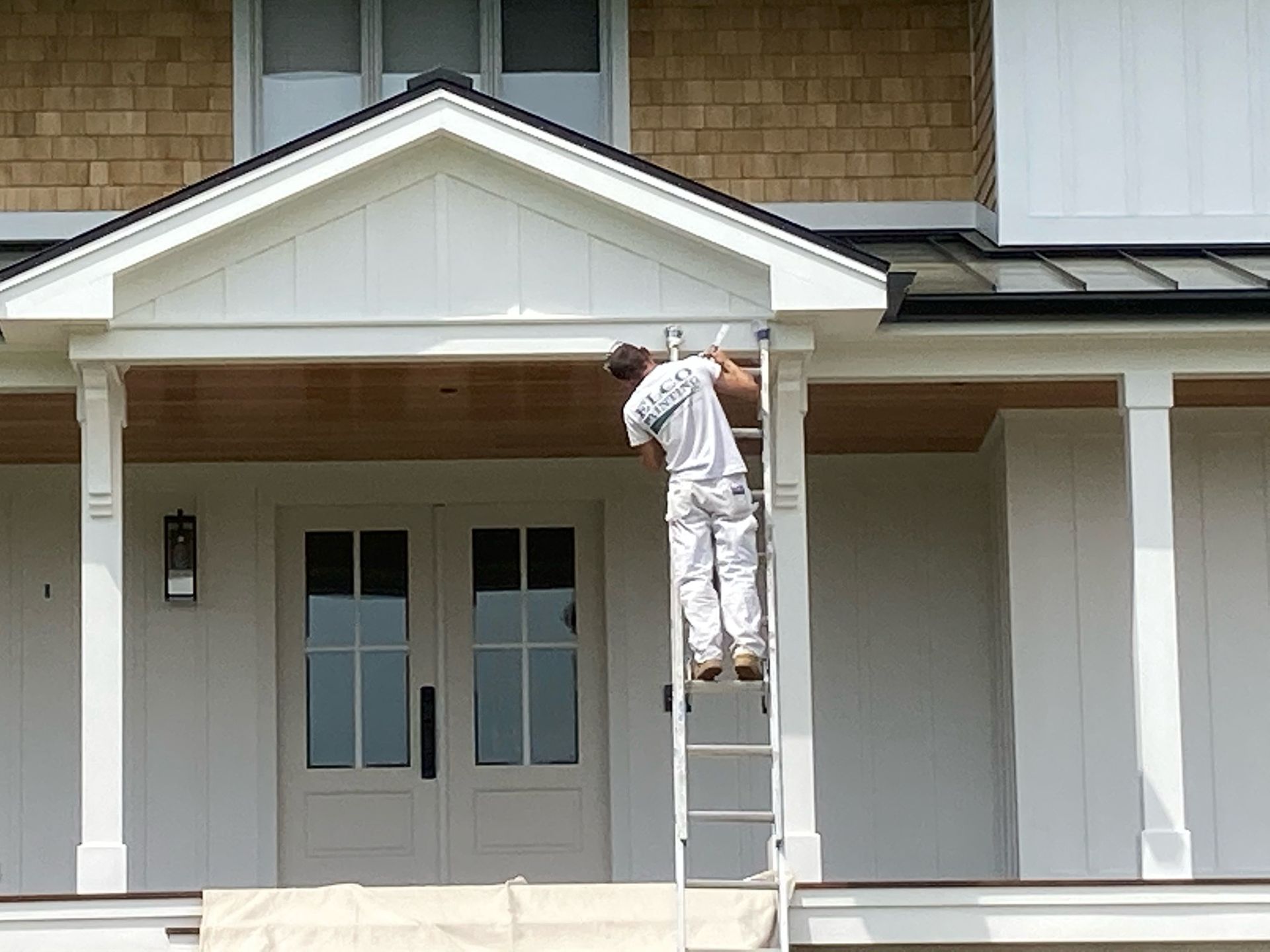 A painter on a ladder is working on the white trim of a house's porch. The house has a light brown facade and a white door.
