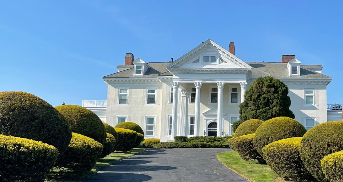 White mansion with large columns and trimmed bushes lining a paved driveway under a clear blue sky.