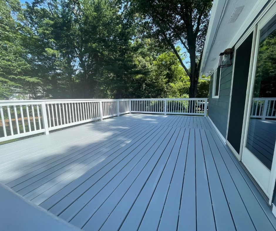 A large, empty deck with gray painted wood and white railings is next to a house with a dark green exterior and sliding glass doors.