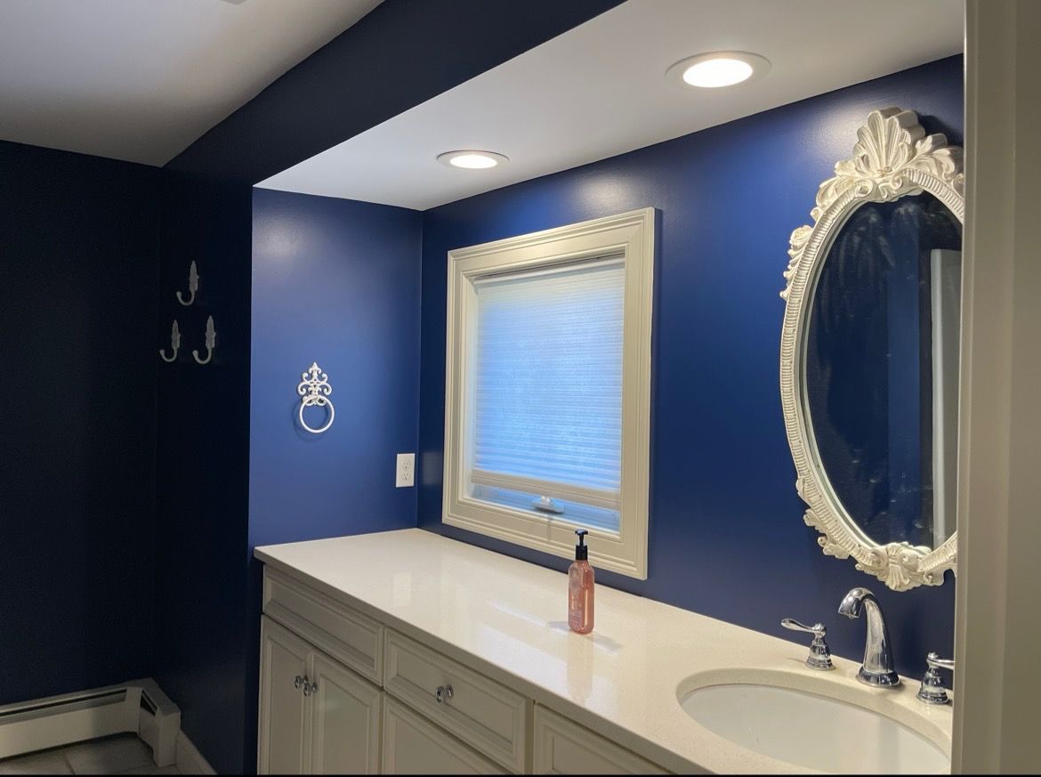 A navy blue bathroom with a white vanity, oval mirror, and window. Three silver hooks hang on the left wall.