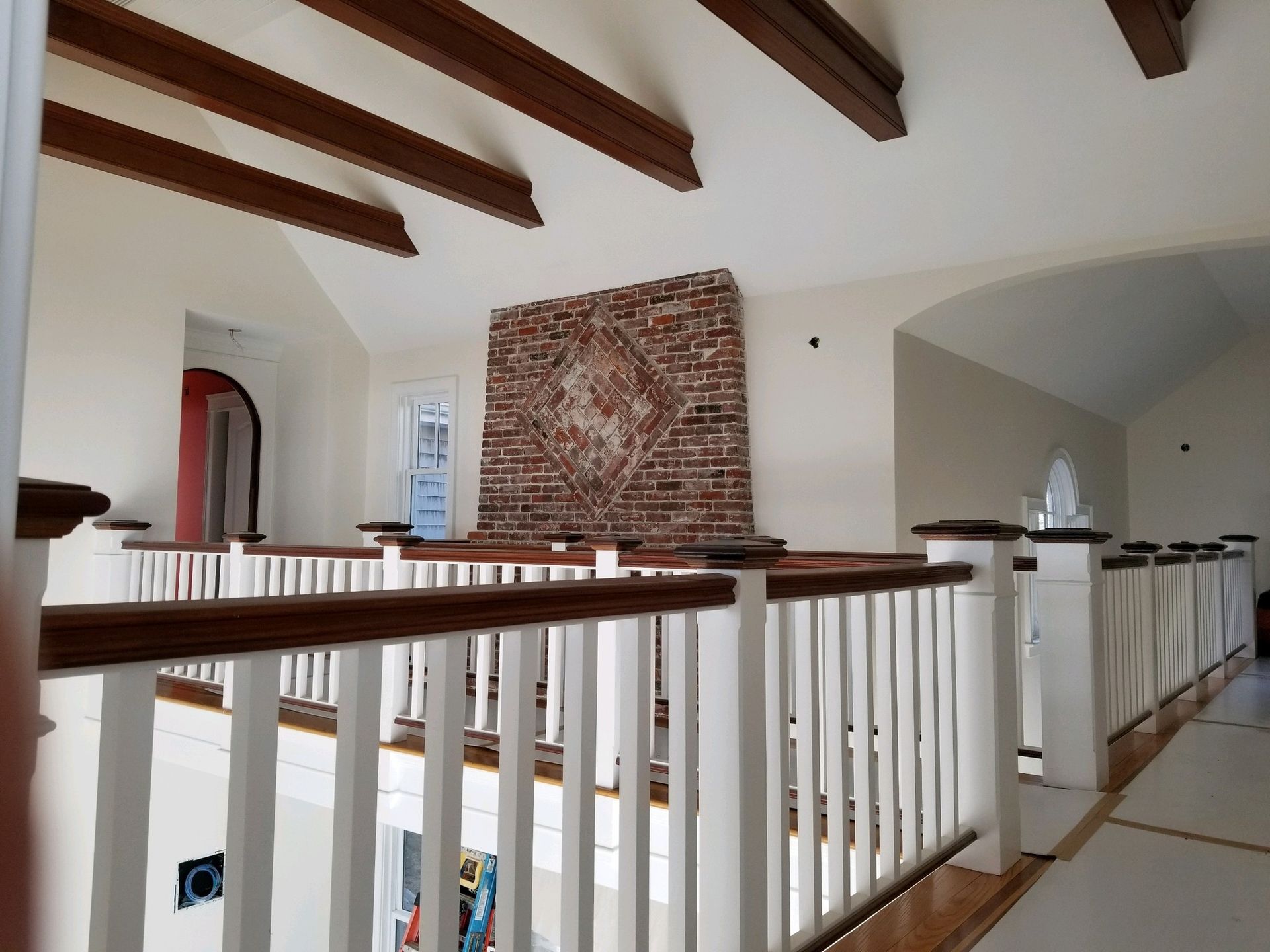 Interior view of a balcony with white railing, brown handrail, and exposed wooden beams. A brick wall forms a backdrop.