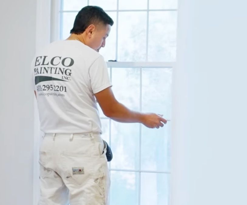 A painter in a white shirt and pants is working on a window inside a room with white walls.