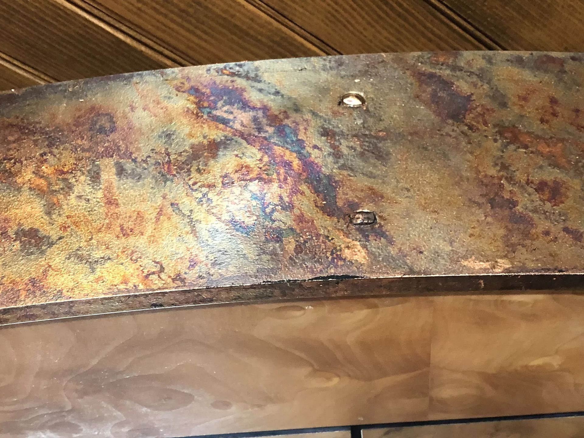 Close-up of a weathered, rusty brown metal beam against a wood-paneled ceiling. The beam has some holes.