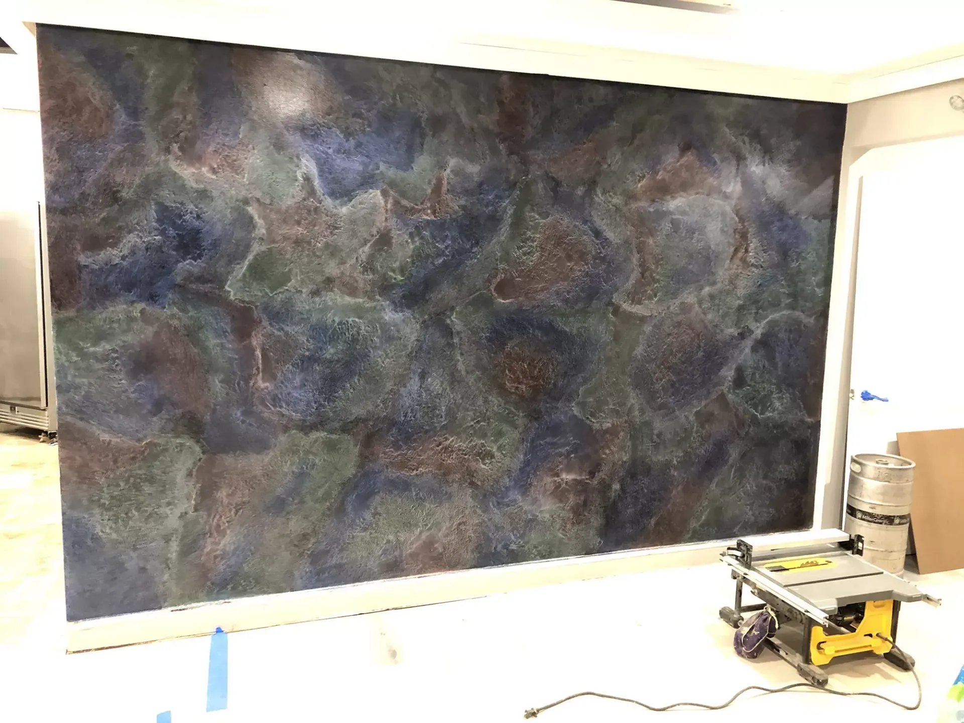 A wall with a dark, textured, mottled paint finish in shades of blue, brown, and green. A saw and other construction materials are on the floor.