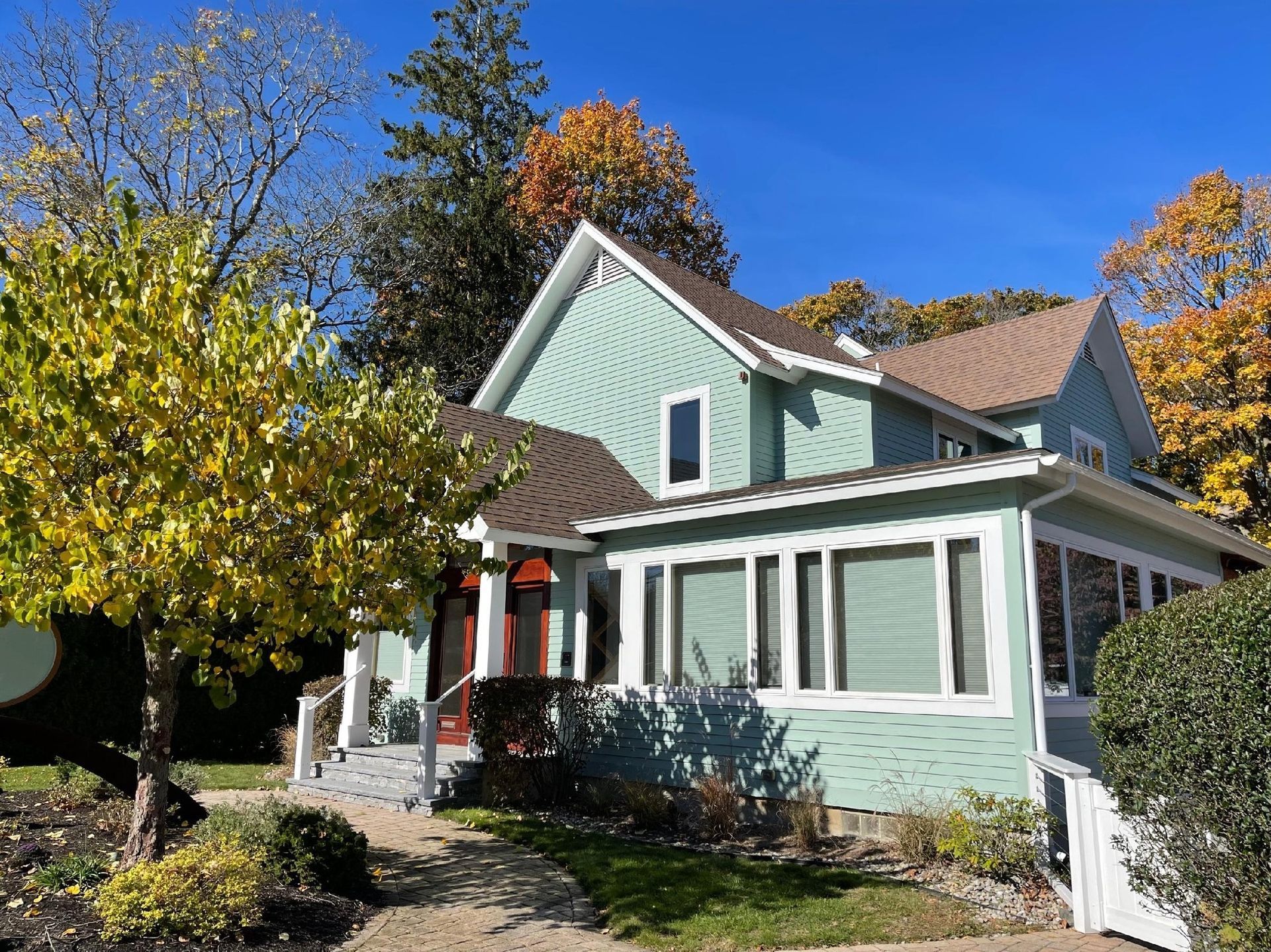 A light green two-story house with brown roof and white trim, with autumn trees and bushes in front.