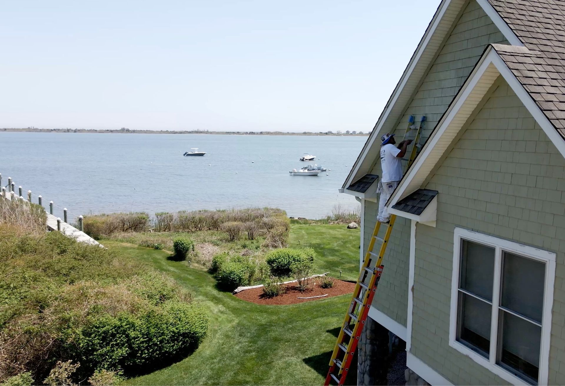 A person paints the side of a light green house, standing on a tall ladder overlooking the water.