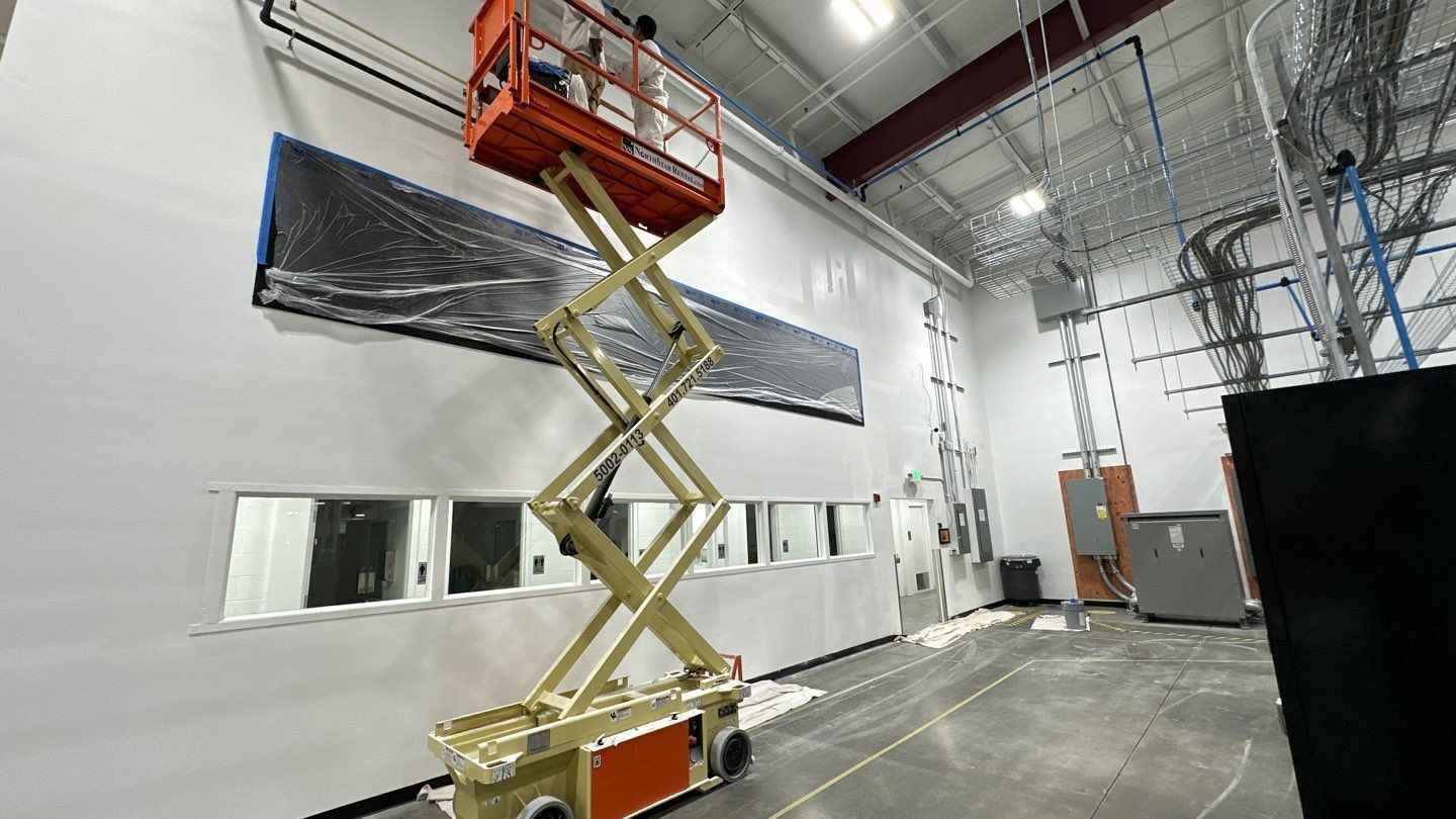 A worker on a scissor lift installing something on a wall inside a building. The wall is white, and there are windows.
