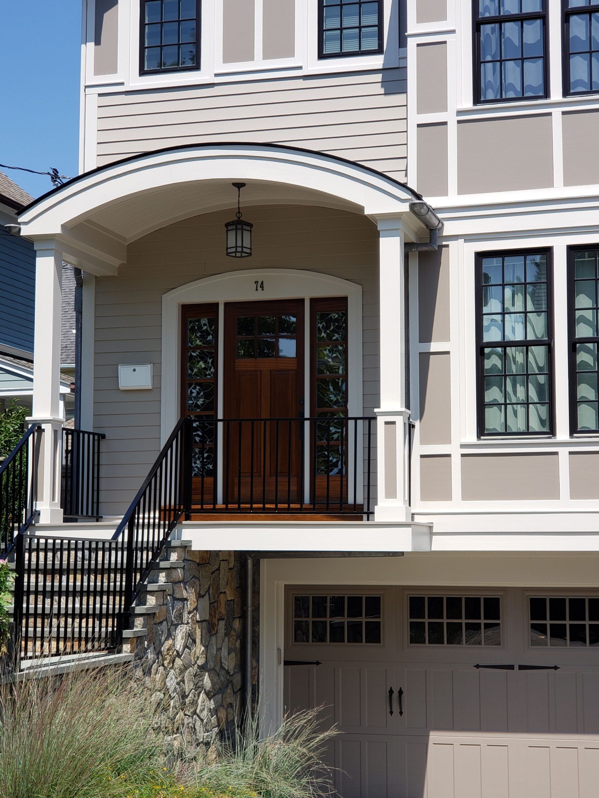 Beige and brown house exterior with arched entryway, steps, and garage door. Black railing and trim, stone foundation.