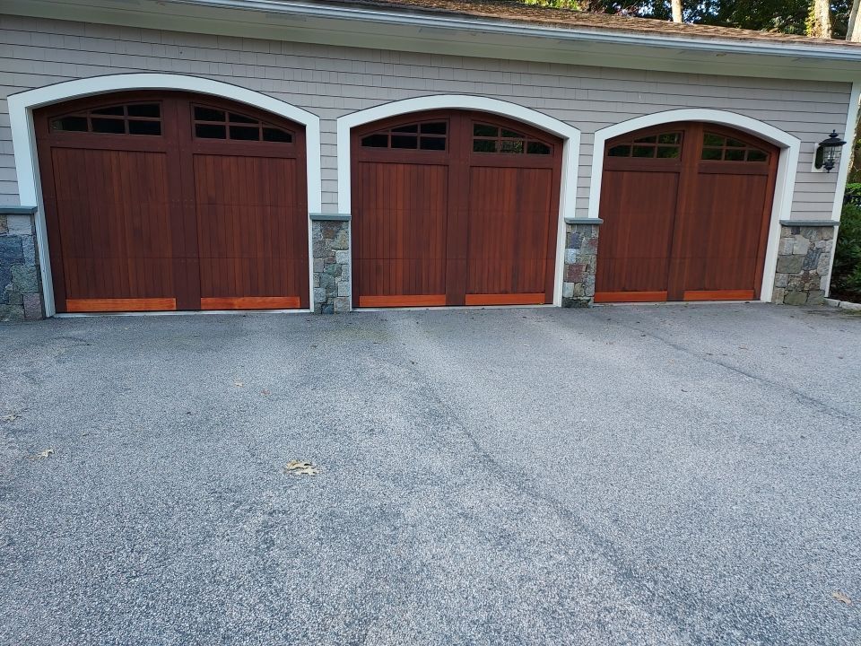 Three wood garage doors with arched tops and stone columns, set in a light-colored building with a dark asphalt driveway.