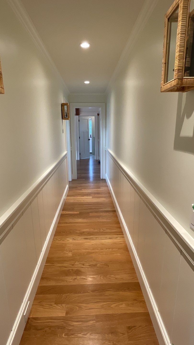A long hallway with wood floors, white walls, and decorative molding. A few doors are visible at the end of the hallway.