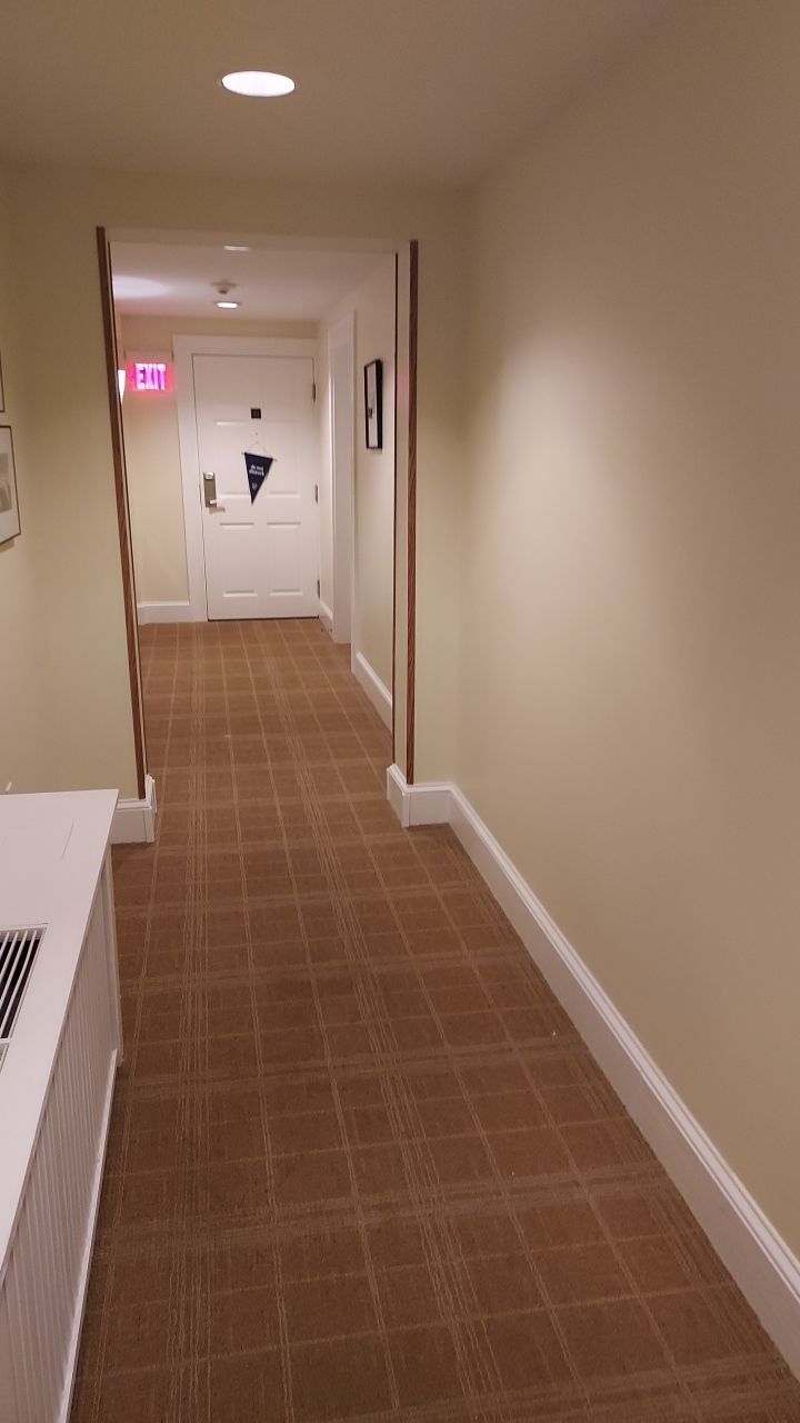 Hallway with brown carpet, cream walls, and a closed door at the end with an illuminated exit sign.