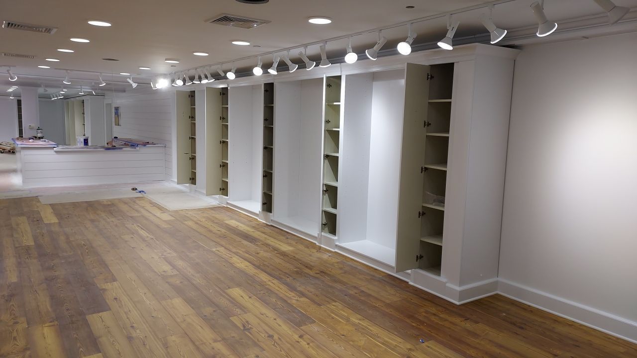 Empty retail space with light-colored wooden flooring, white walls, and built-in shelving. Overhead track lighting illuminates the area.