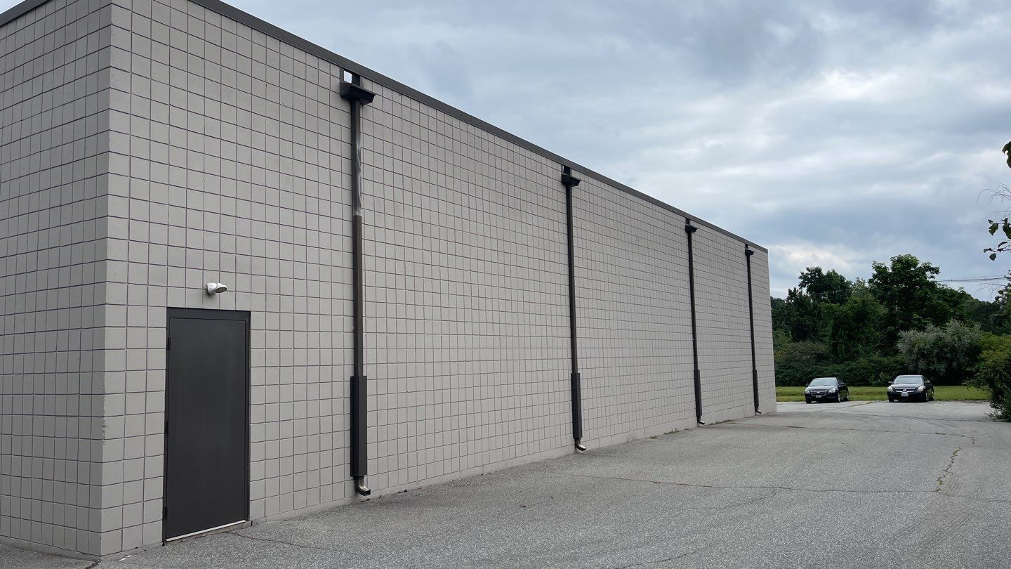 Exterior of a large, gray industrial building with a single dark door. Two cars are parked in the gravel lot.