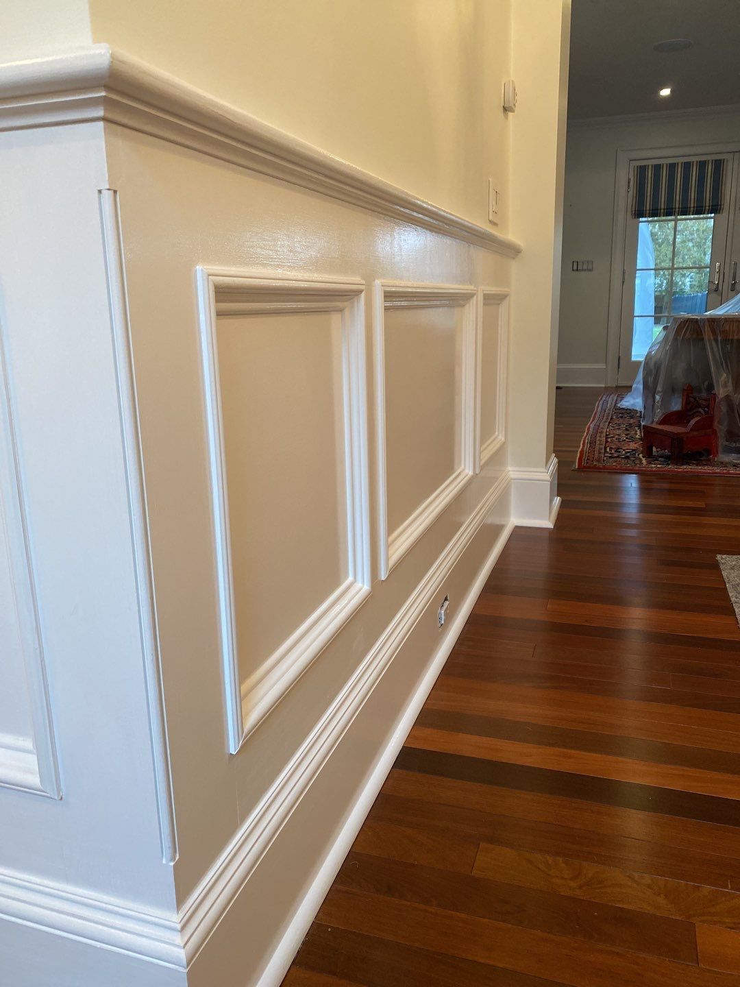 Beige wall with white trim, wainscoting, and dark wood flooring. A doorway leads to a brightly lit room.