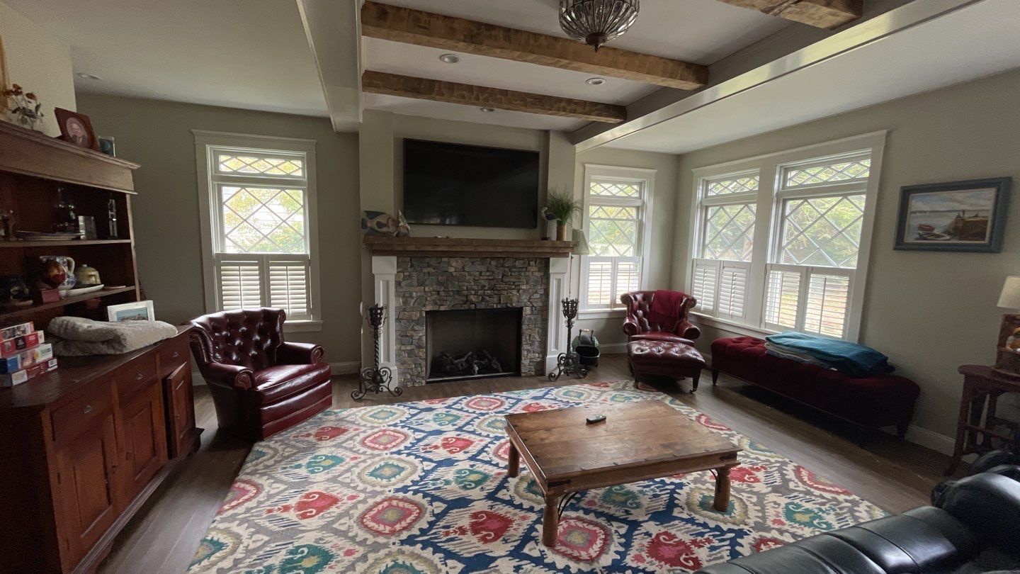 Living room with a stone fireplace, TV, red leather chairs, and a patterned rug. Large windows with light coming in.