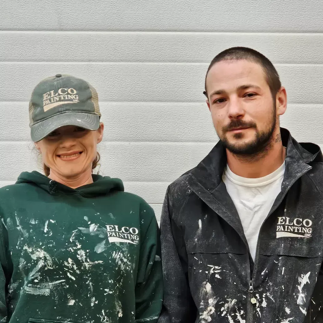 Two people, a woman and a man, smiling at the camera. Both are wearing work clothes covered in paint, with logos that read 