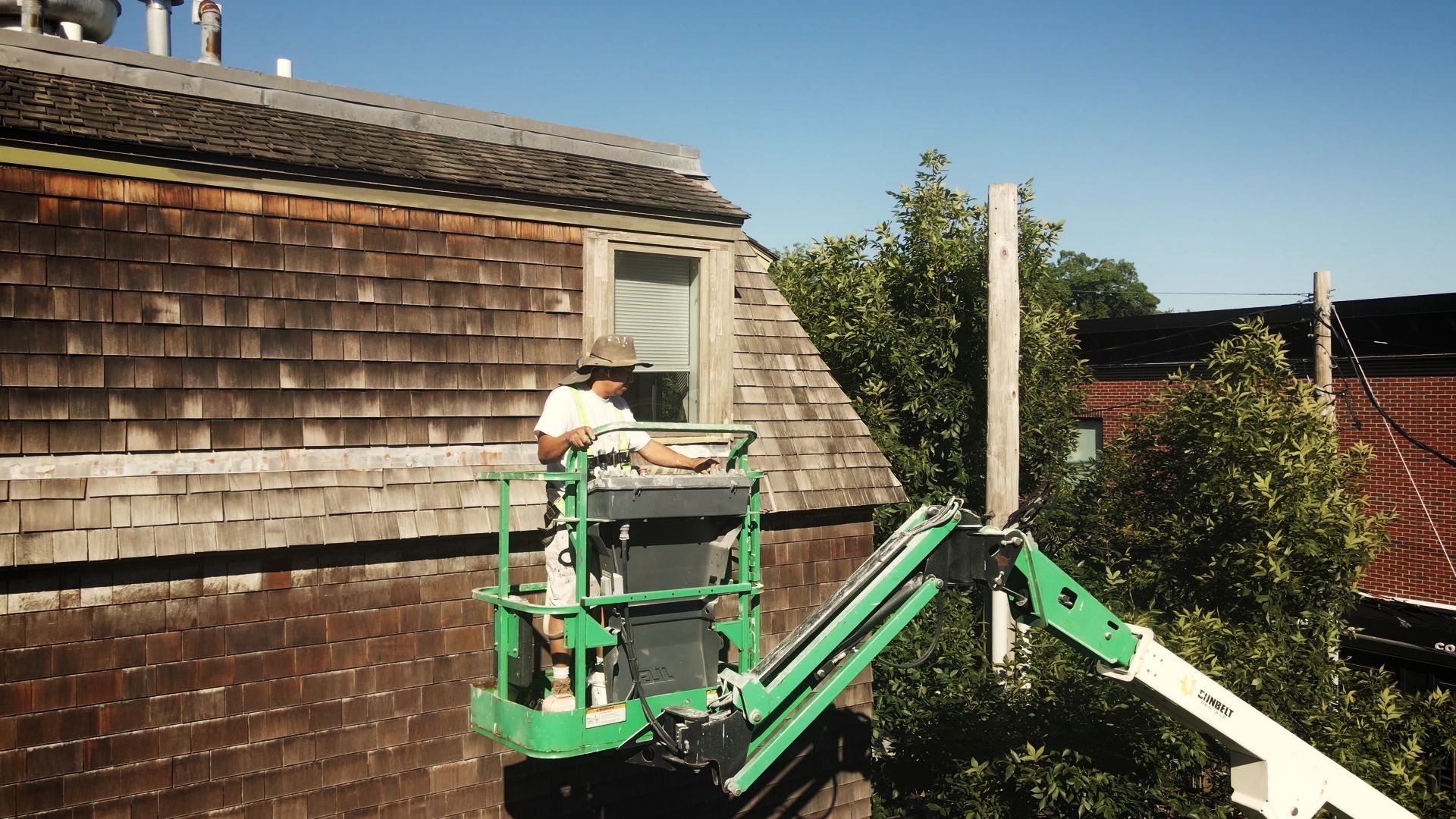 A worker repairs a building's exterior from a green lift. The building has a weathered brown shingle roof.